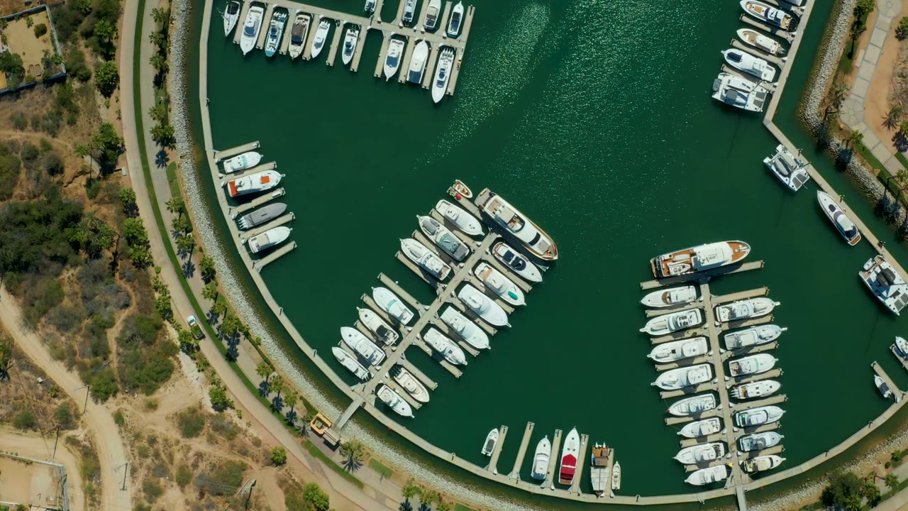 Mid-early frame over Puerto Los Cabos marina showing a broad green basin, packed white slips, a few red details on boats, shoreline paths, and no visible people.