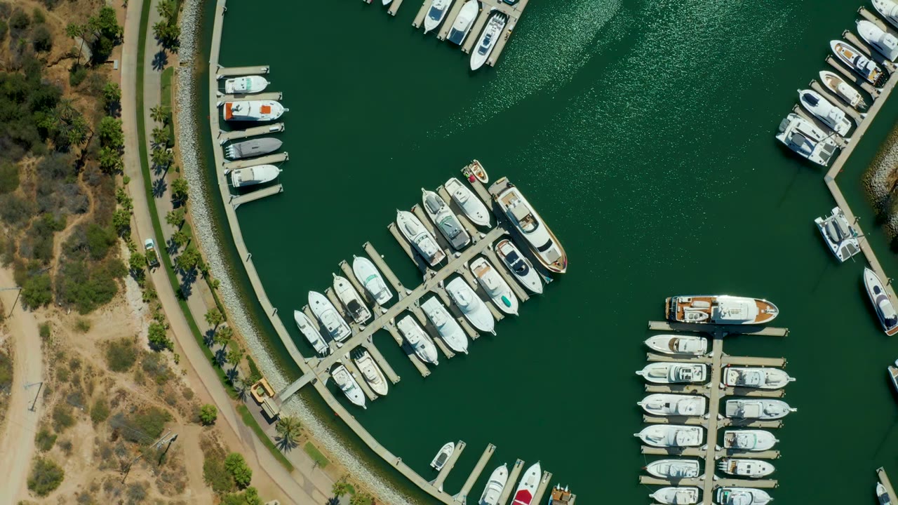 Overhead view of the Puerto Los Cabos marina showing docked white boats, green rippled water, curved shoreline paths, dry scrub land, and no visible people.