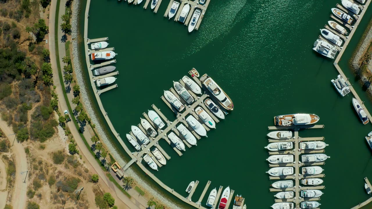 Top-down Puerto Los Cabos marina view with the moving boat shifted across the open water, rows of white docked vessels around the slips, dry shoreline terrain, and no visible people.