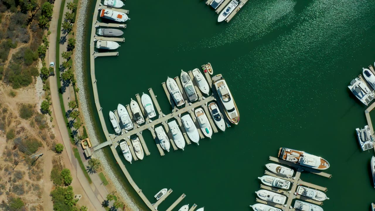 Overhead view of Puerto Los Cabos marina showing rows of docked white boats, dark green water, a curved shoreline road, dry brush, and no visible people at the start of the clip.