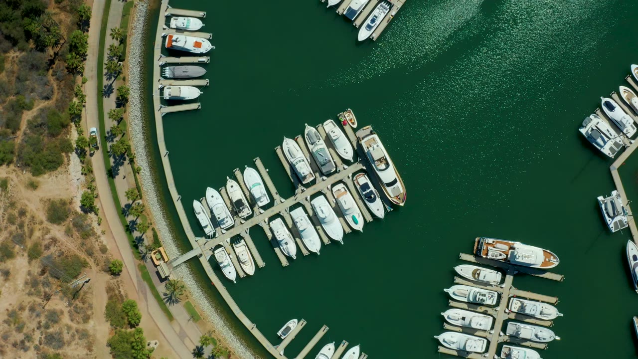 Top-down Puerto Los Cabos marina view with white yachts lining docks, a small boat near the lower center, green water texture, dry shoreline terrain, and no visible people.