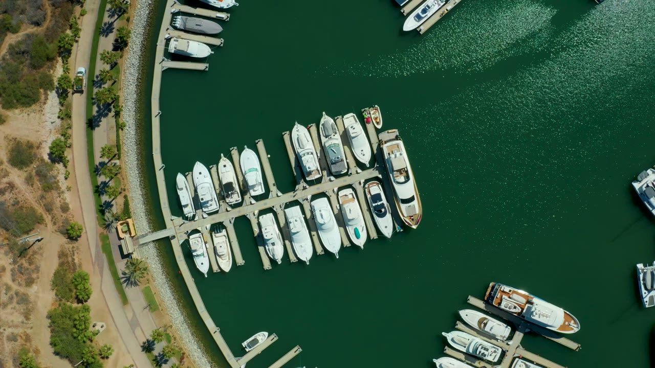 Overhead Puerto Los Cabos marina frame with docked yachts grouped around a pier, a shoreline road and scrubland along the left side, and no visible people.