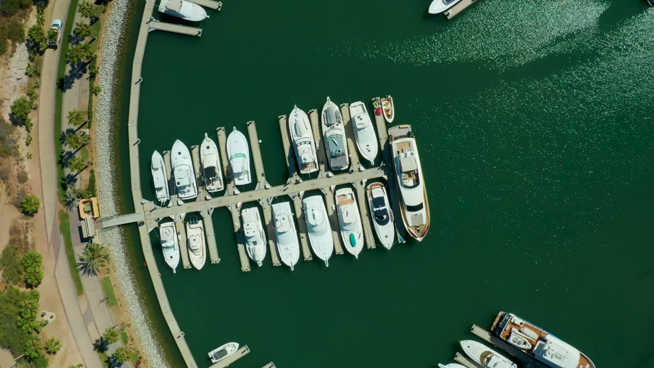 Top-down view of the Puerto Los Cabos marina with the dock and boats shifted left in frame, open green water on the right, a vessel edge near the lower right, and no visible people.