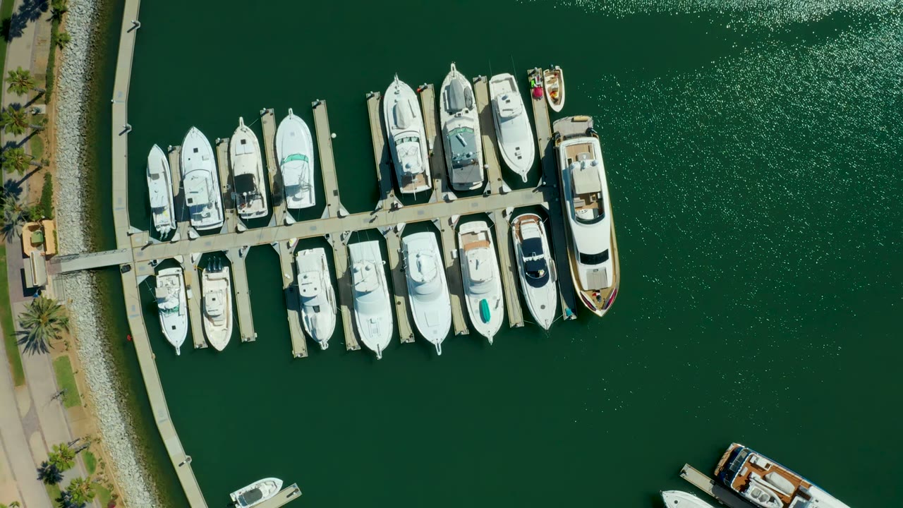 Top-down frame over Puerto Los Cabos marina showing white yachts tied to a floating dock, green water, a curved waterfront path at left, and no visible people.