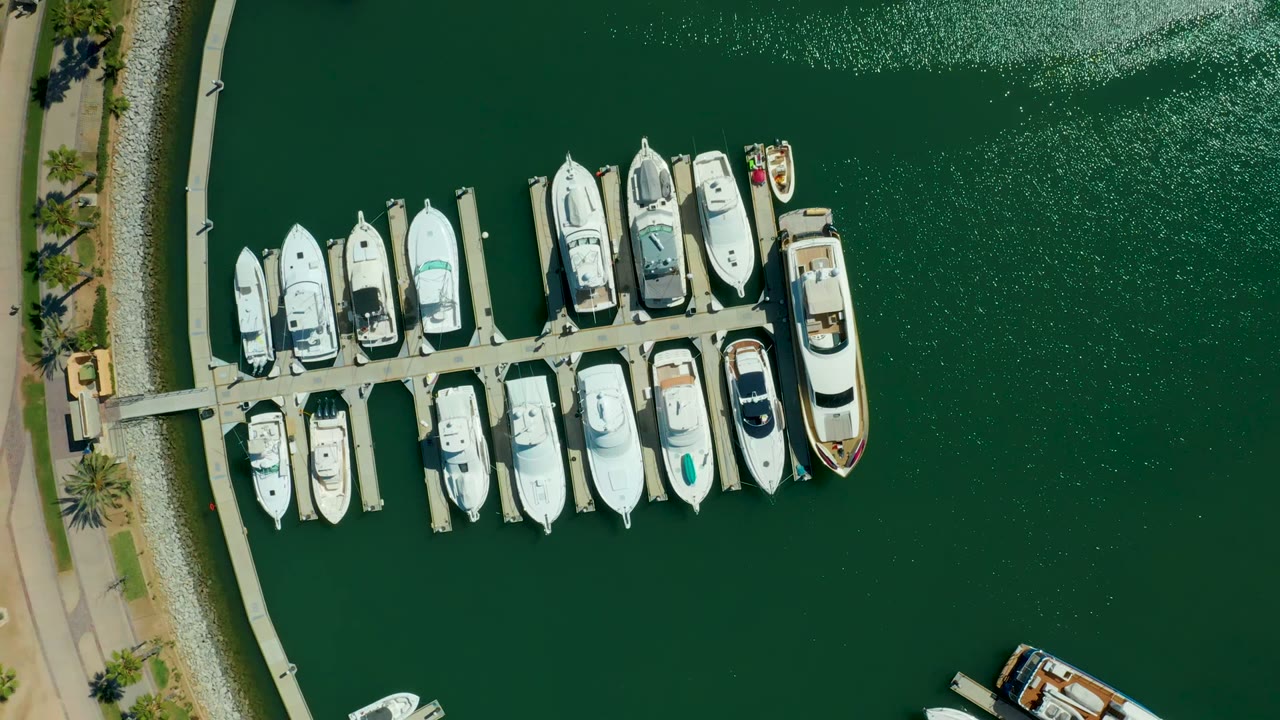 Overhead view in Puerto Los Cabos shows the docked yachts shifted slightly within frame, open green water on the right, the shore path on the left, and no visible people.