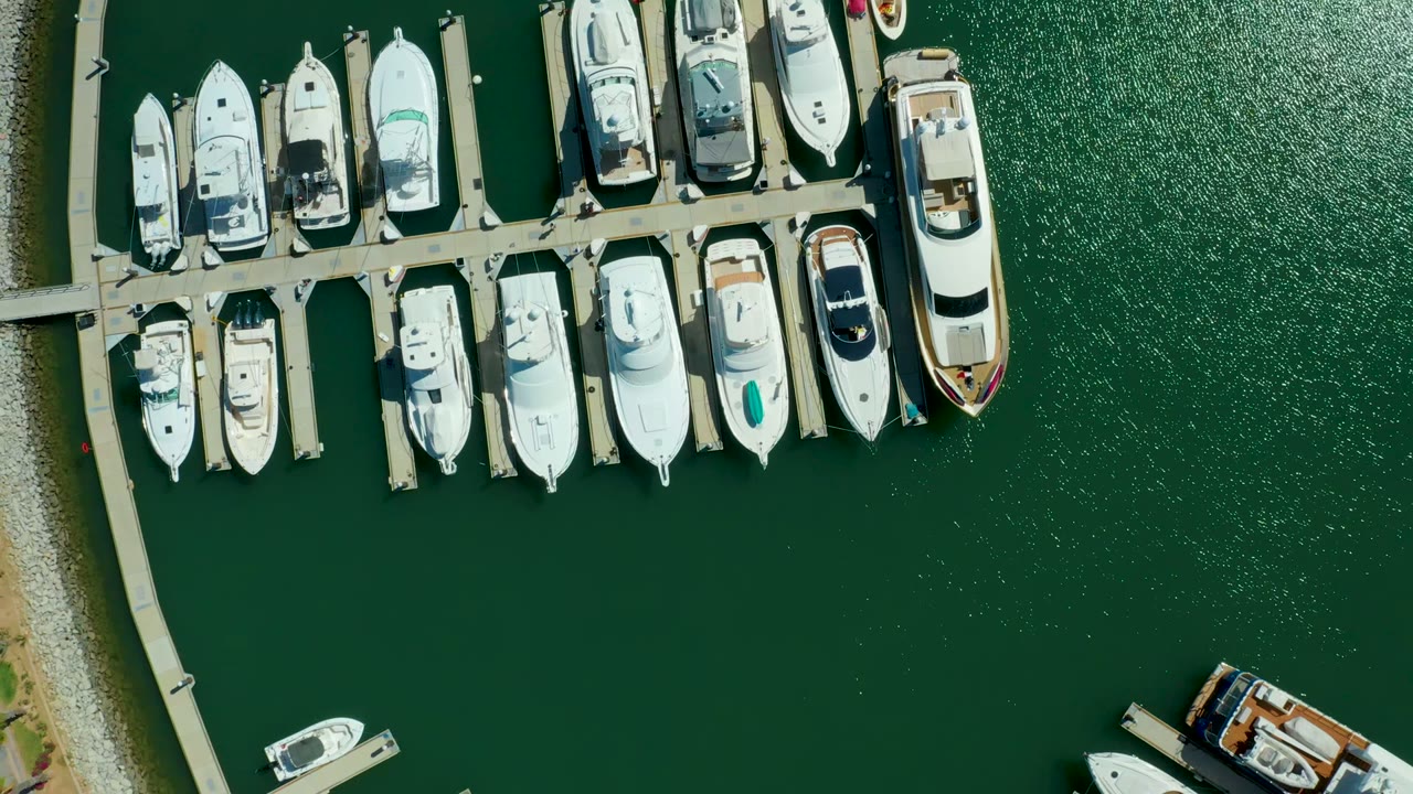 Top-down view of Puerto Los Cabos marina with white boats docked along piers, green water, a curved pebbled shoreline at left, and no visible people.