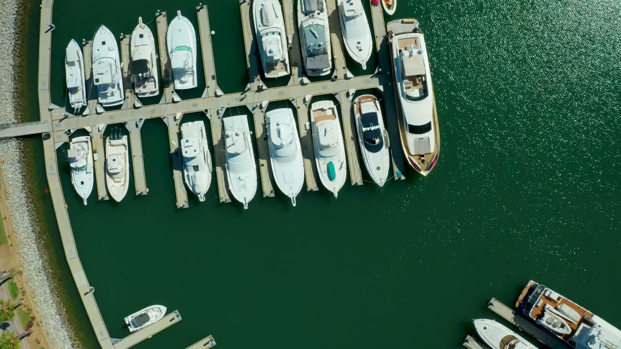 Overhead Puerto Los Cabos marina frame with rows of white boats at the top, open green water through the center, shoreline stones at left, and no visible people.