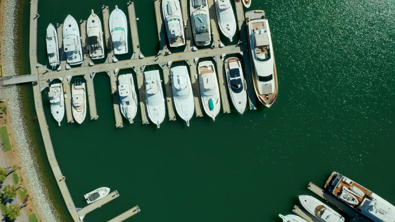 Aerial overhead view in Puerto Los Cabos with docked yachts across the upper frame, bright green marina water, a narrow shoreline curve, and no visible people.