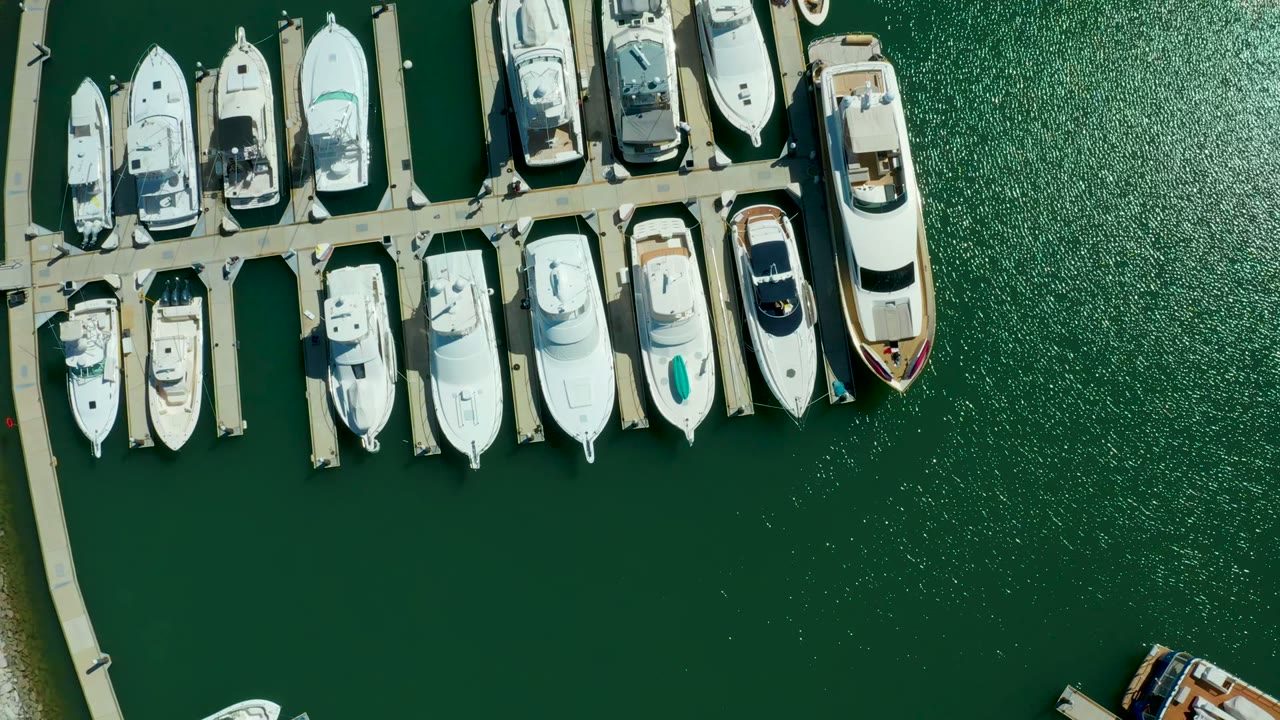 Aerial overhead view in Puerto Los Cabos showing parallel slips packed with white boats, open green water to the right, and no visible people.