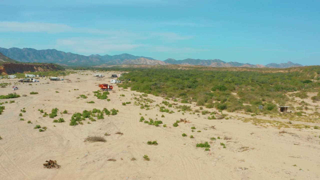 Wide aerial view in Los Frailes Los Cabos, Baja California Sur showing sandy terrain, scattered shrubs, distant mountains, a small red truck at mid-left, and no visible people.