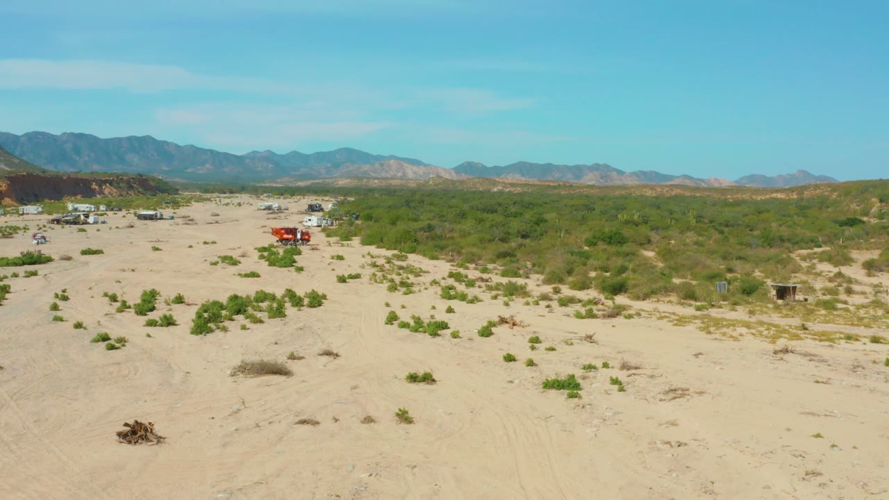 Red truck crossing open sand with shrubs and distant mountains.