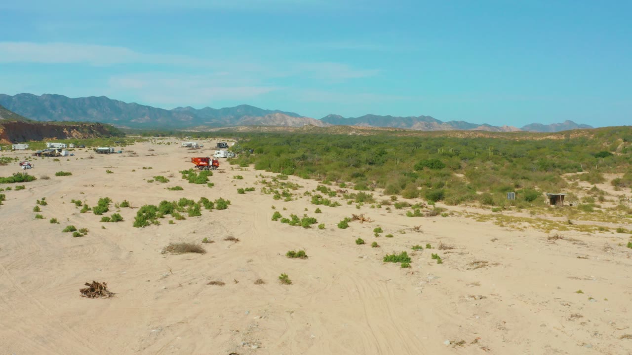 Wide drone-like view over Los Frailes Los Cabos arid sand and scrub, with the red truck in the midground near a cluster of pale vehicles or structures and no visible people.
