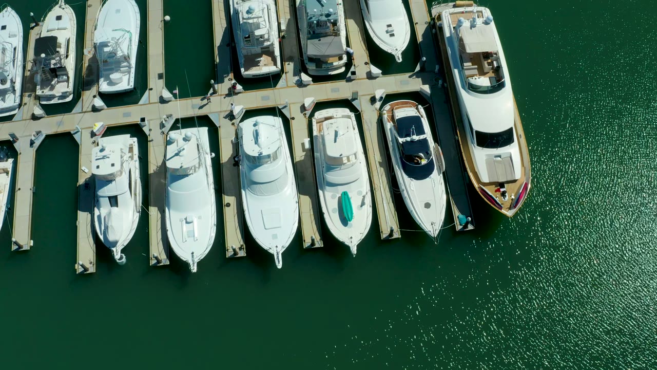 Aerial overhead frame in Puerto Los Cabos with docked yachts clustered near the top edge and textured green water filling the lower frame, with no visible people.