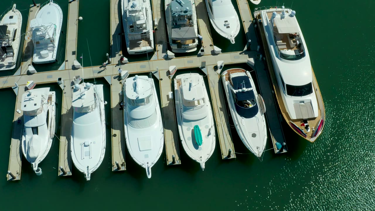 Puerto Los Cabos marina boats remain aligned in a top-down composition, with a large white yacht at the right edge and textured green water to the side, no visible people.