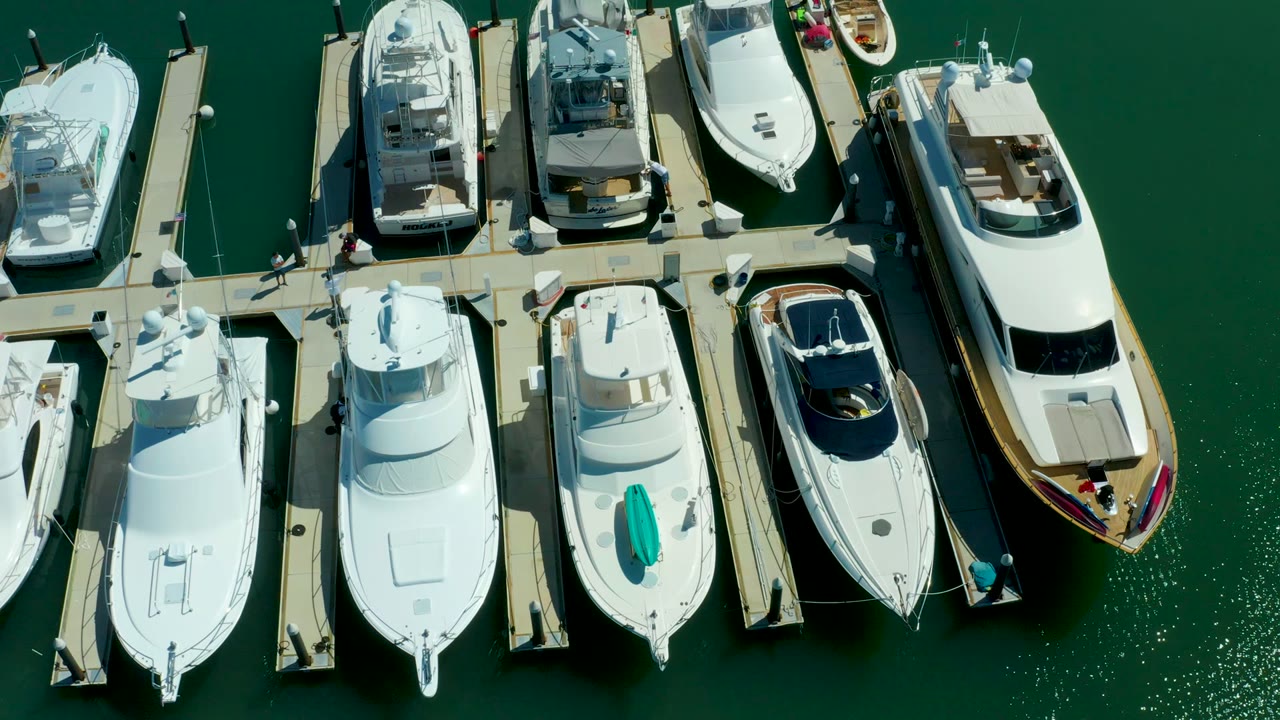Overhead view of docked yachts and narrow piers in Puerto Los Cabos, with green marina water and tiny people visible on the docks.
