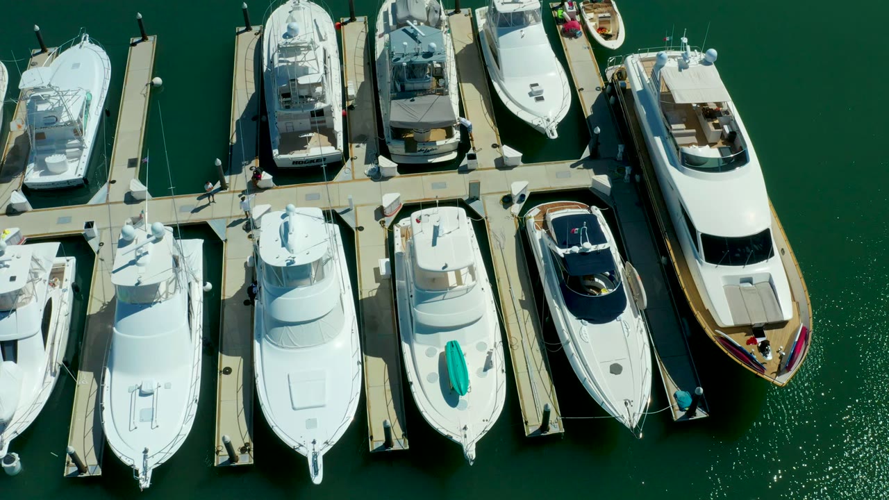 Top-down view in Puerto Los Cabos with white yachts moored beside straight piers, calm green water, and small figures visible on the dock.