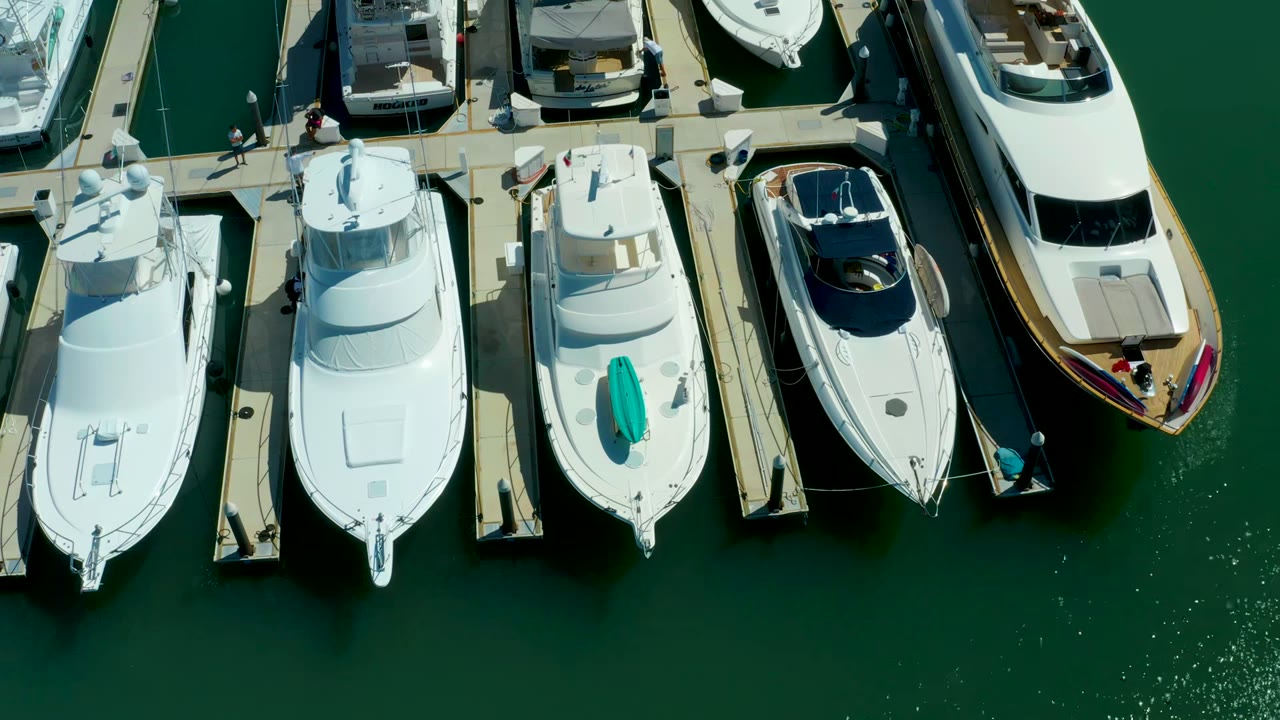 Overhead view of white yachts and small boats docked in green marina water at Puerto Los Cabos, with narrow piers and small people visible near the upper dock.