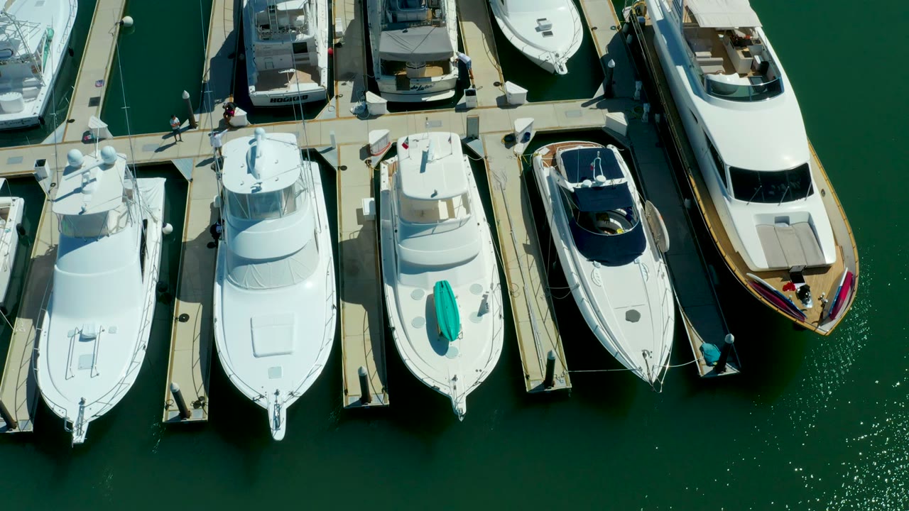 Top-down view at Puerto Los Cabos with a row of white yachts, a larger vessel at the right edge, bright dock surfaces, and sun sparkle on green water.
