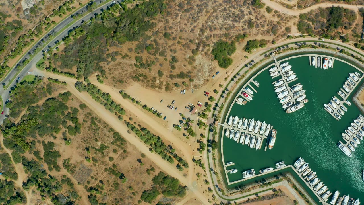 Opening overhead frame of Puerto Los Cabos marina with green water, white boats on docks, a curved road, sandy parking area, dry scrubland, scattered trees, and no visible people.