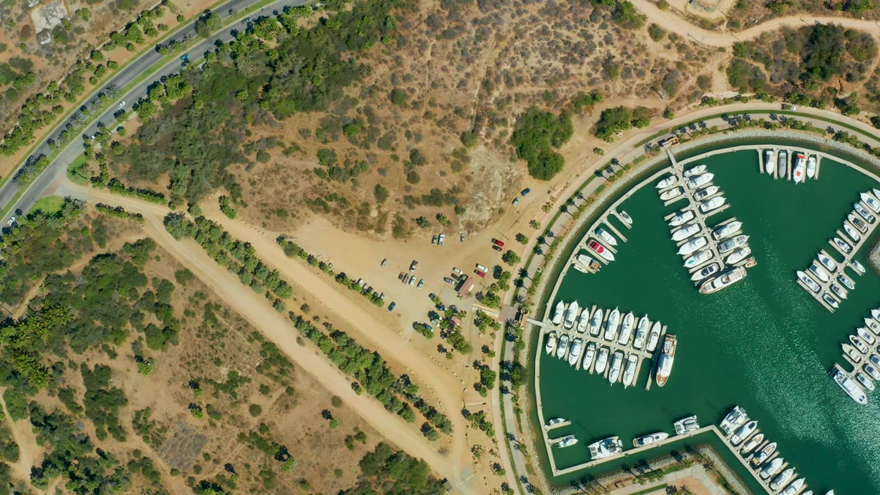 Overhead view of Puerto Los Cabos marina with green water, rows of white boats, a sandy parking area, curving roads, dry scrubland, and no visible people at the start of the clip.
