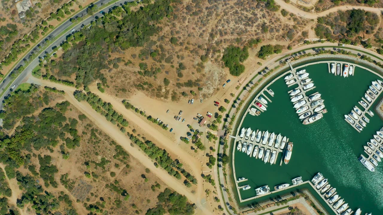 Top-down view in Puerto Los Cabos with clustered boats in marina slips, green water, a curved waterfront edge, sandy vehicle areas, access roads, dry vegetation, and no visible people.
