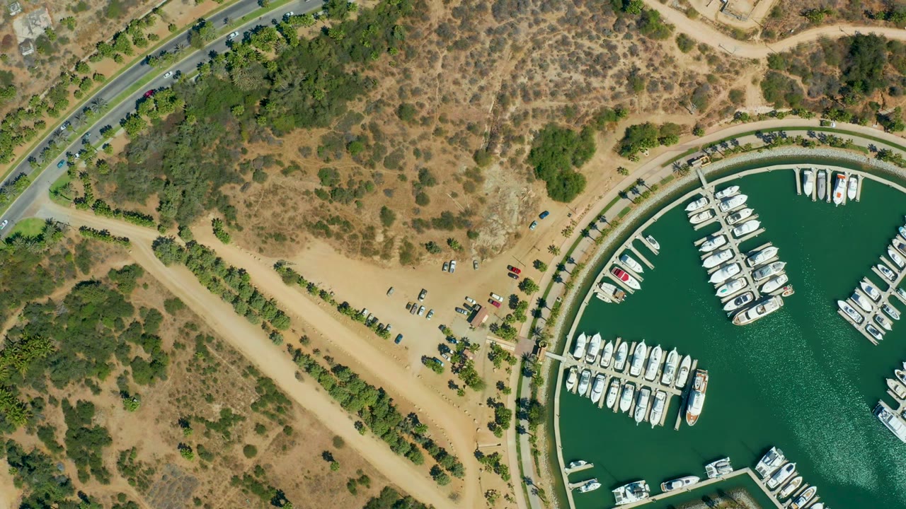 Overhead view of Puerto Los Cabos marina with white boats along docks, green-blue water, a curved road, sandy parking area, dry brush terrain, and no visible people at 0:00.