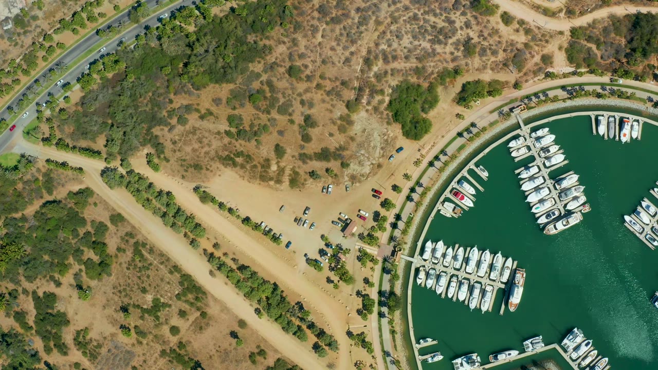 Overhead view of Puerto Los Cabos marina with white boats in green water, a curved road at left, sandy parking areas, dry hills, and no visible people.