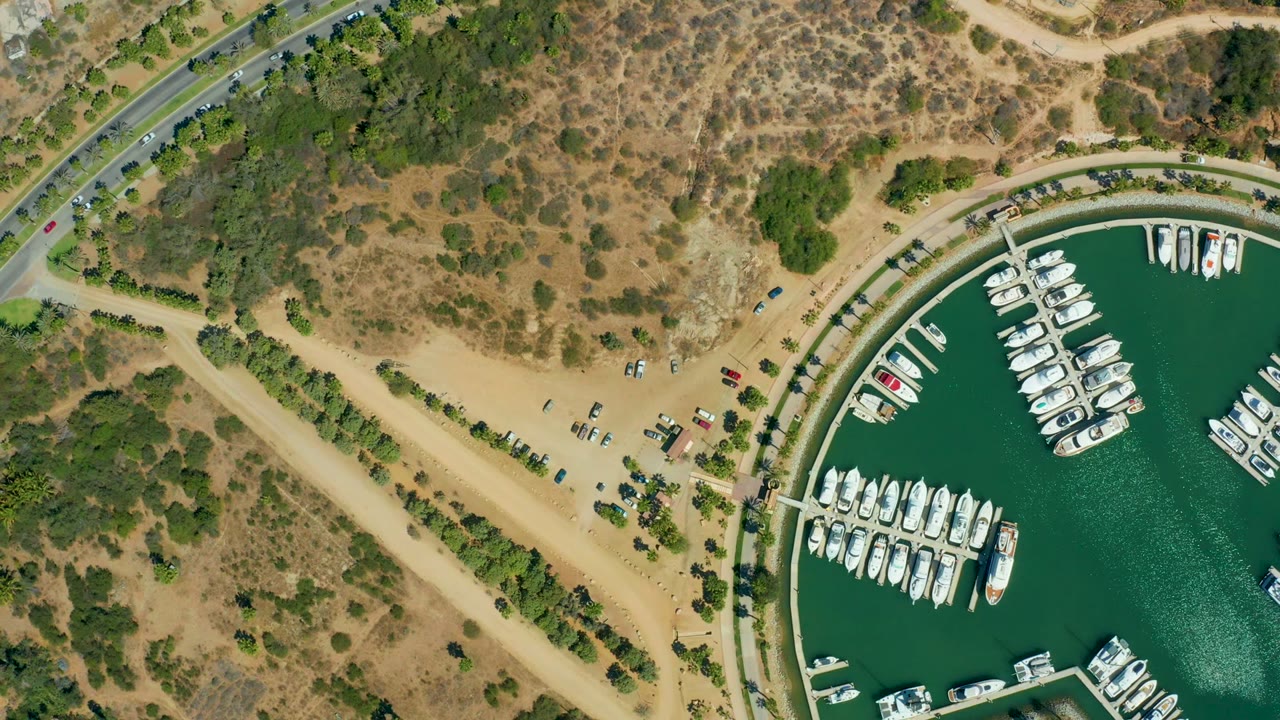 Aerial view of dock rows, green marina water, road traffic, and arid terrain.