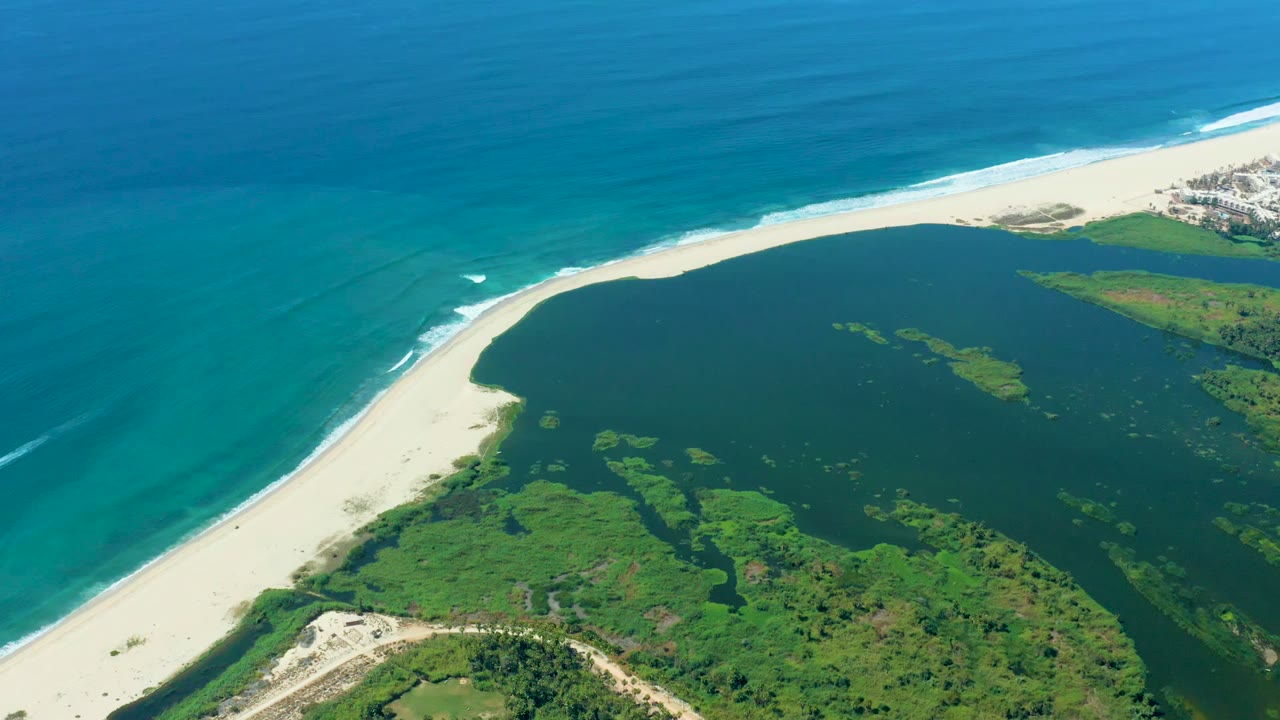 High aerial coastal view with ocean, curved beach, lagoon, greenery, and distant development.