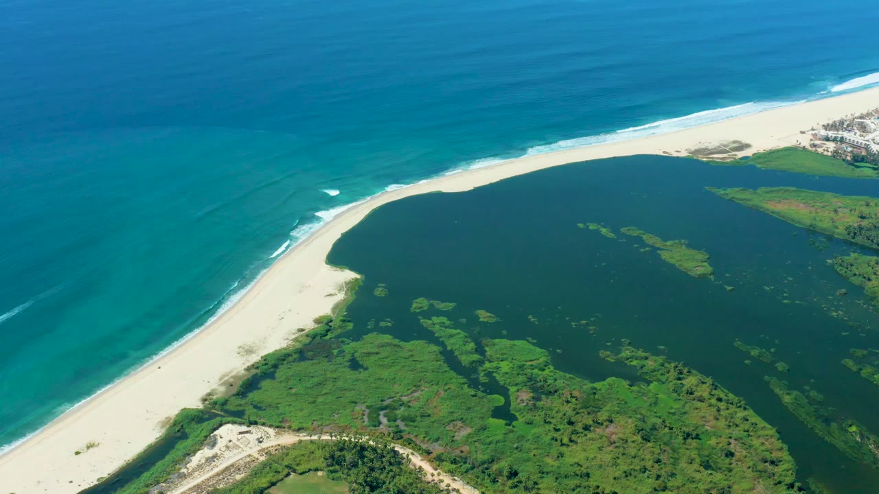 A wide drone-style frame over Puerto Los Cabos shows pale sand separating the turquoise ocean from the darker lagoon, with green vegetation patterns inland and no visible people.