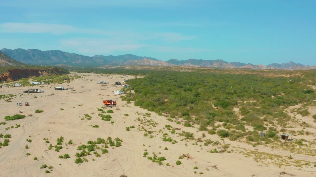 Aerial frame over Los Frailes desert showing a red truck, white trailer, scattered vehicles, sandy ground, green shrubs, distant mountains, and no visible people.