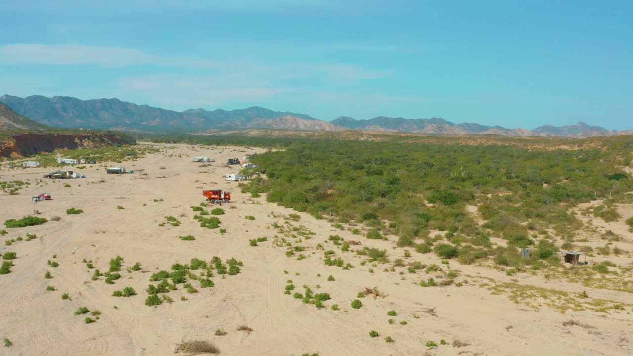 The Los Frailes desert remains in a high wide composition, with vehicles spaced across the sandy clearing, dense green scrub to the right, mountains beyond, and no visible people.