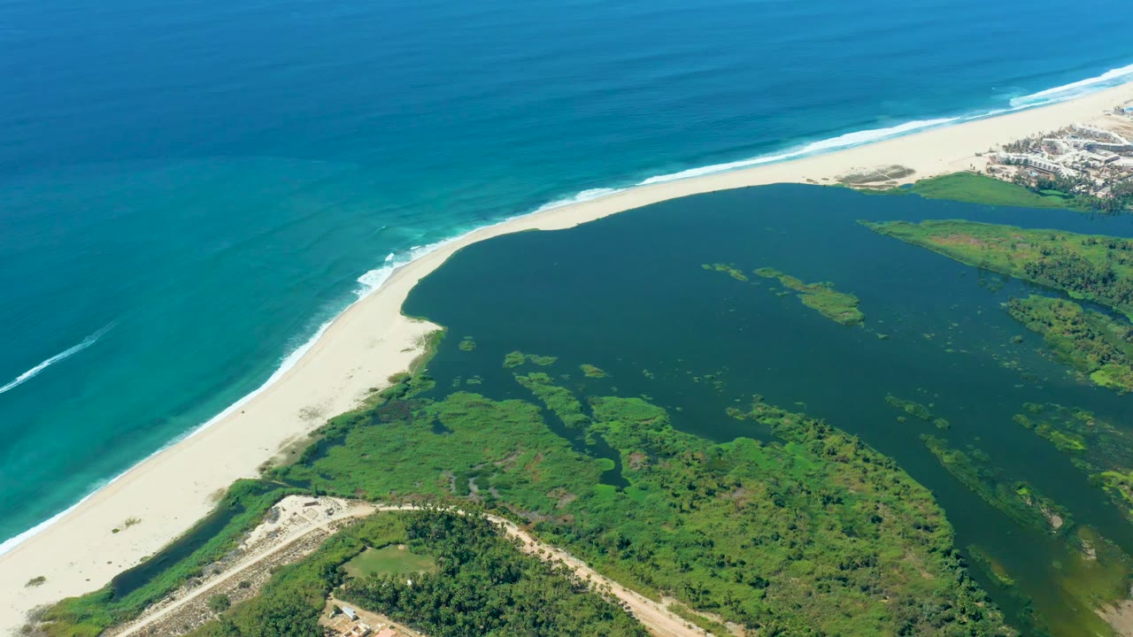 Aerial view over Puerto Los Cabos with turquoise ocean above a white sand beach, a dark lagoon, green vegetation, and a small settlement at the upper right, with no visible people.