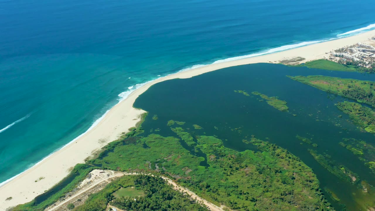 High aerial view in Puerto Los Cabos with dark lagoon water centered below the white beach, turquoise ocean beyond it, green vegetation at the lower edge, distant buildings at upper right, and no visible people.