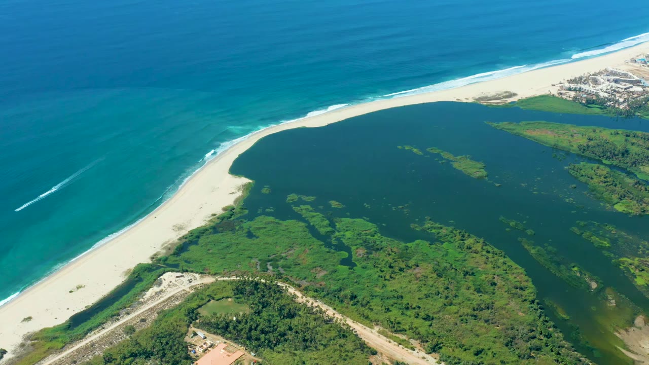 Aerial frame over Puerto Los Cabos showing turquoise ocean, white beach, dark green lagoon, vegetation, a road, scattered shore buildings, and a small boat wake at left with no visible people.