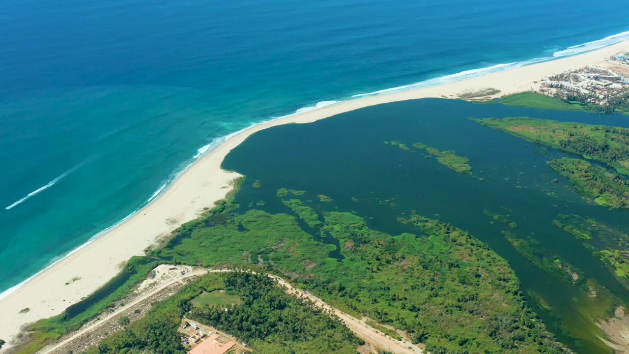 Aerial frame from Puerto Los Cabos showing the blue ocean meeting a bright sand beach, the lagoon spreading inland, green vegetation and a road in the lower frame, and no visible people.