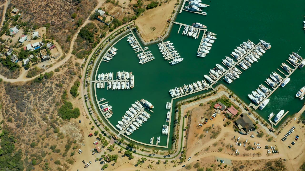 Mid-clip overhead frame of Puerto Los Cabos marina with packed slips, dark green water, tan shoreline roads, parked vehicles, dry brush, and no visible people.