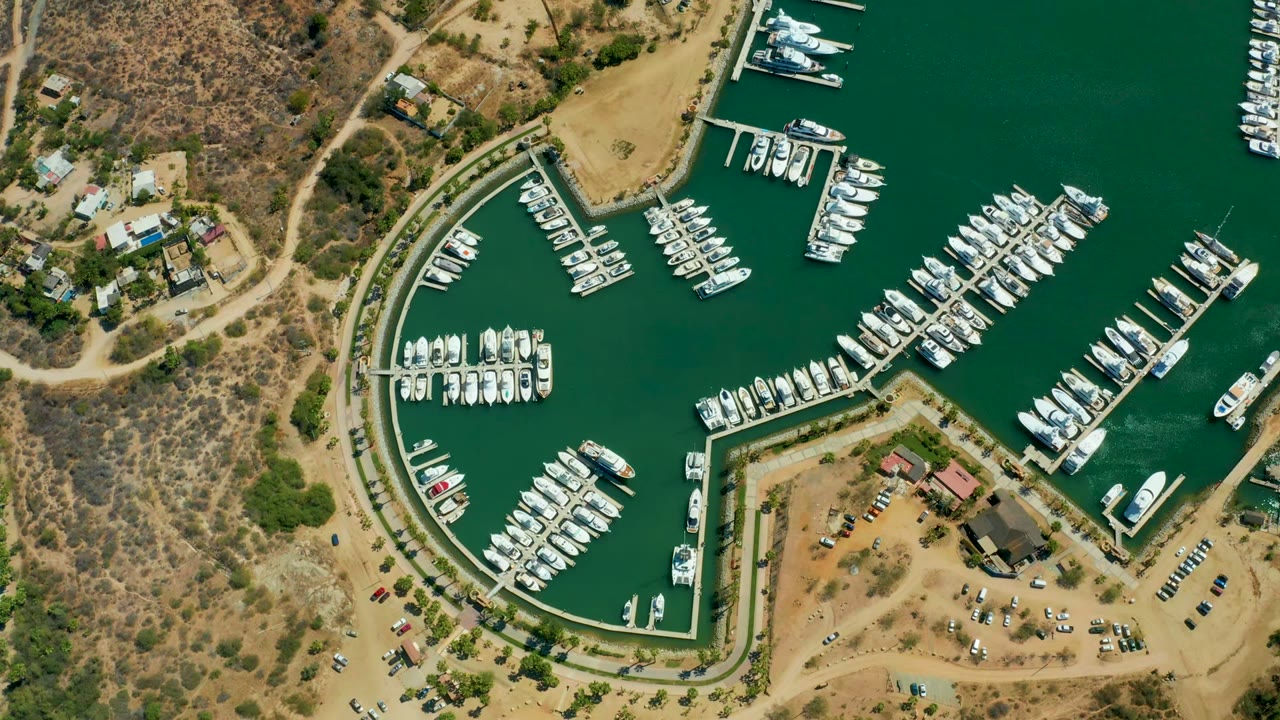 High drone view over Puerto Los Cabos with yacht slips, green harbor water, tan dry land, roadside parking, small buildings, and no visible people in the frame.