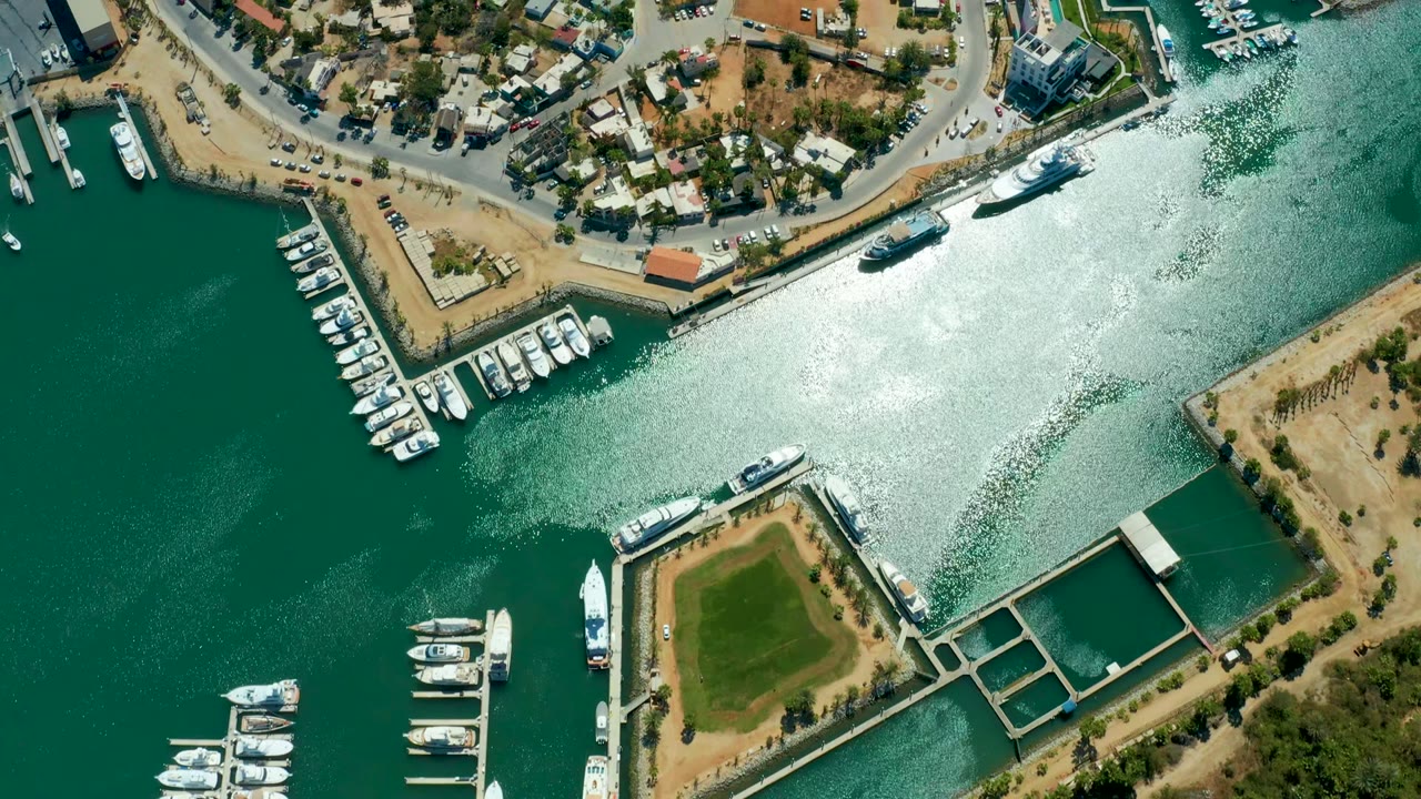 Aerial view over Puerto Los Cabos marina water with docked white boats, waterfront roads, dry shoreline, bright sun glare on the channel, and no visible people.
