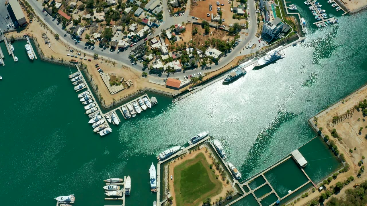 Puerto Los Cabos marina from above, with a cluster of white yachts at docks, a boat progressing through blue-green water, strong reflected light, and no visible people.