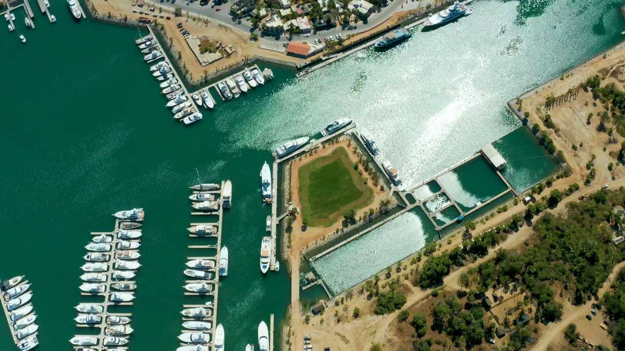 Puerto Los Cabos aerial frame at the start of the clip shows marina docks lined with white boats, green water, bright channel glare, shoreline buildings, trees, parking areas, and no visible people.