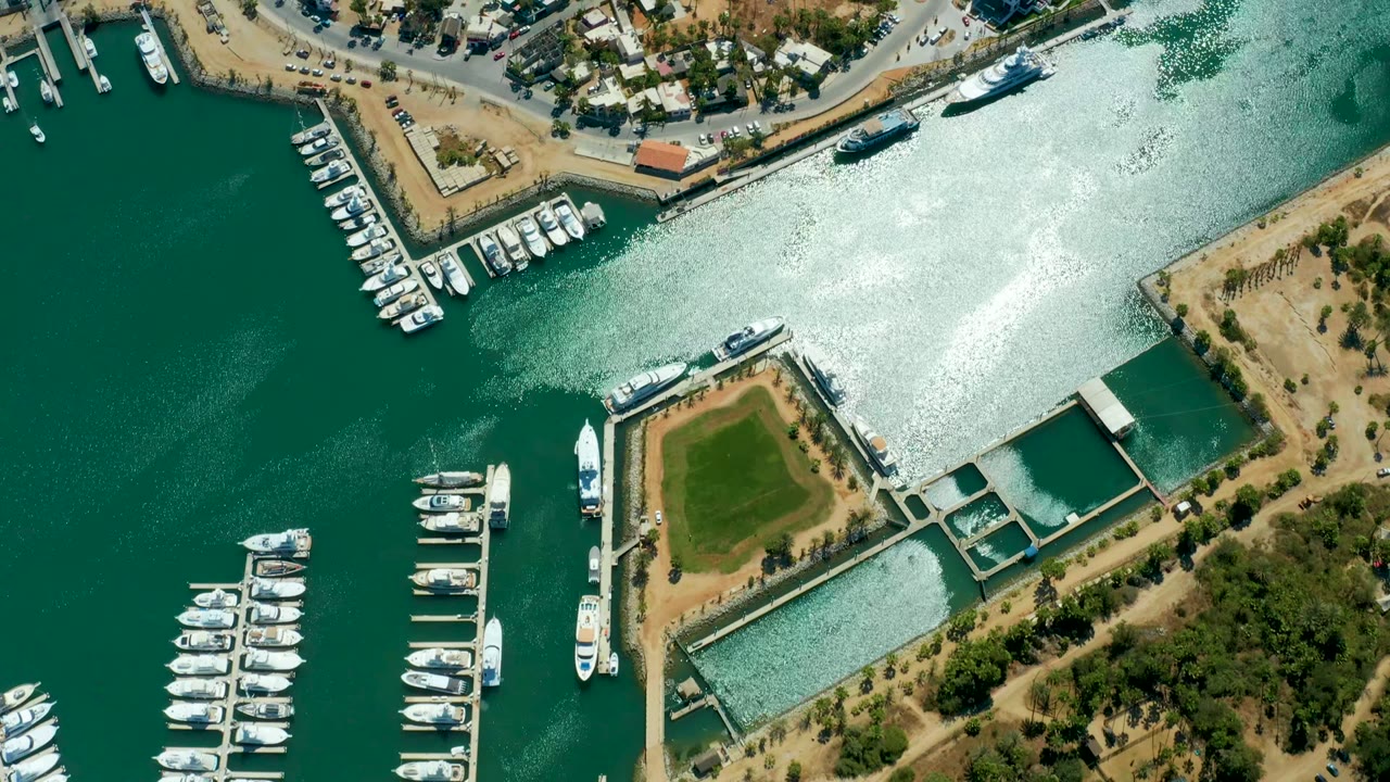 At 0:02, the aerial view shows Puerto Los Cabos docks with rows of boats, a small moving wake in the left water, bright glare across the upper channel, shore lots, trees, and no visible people.