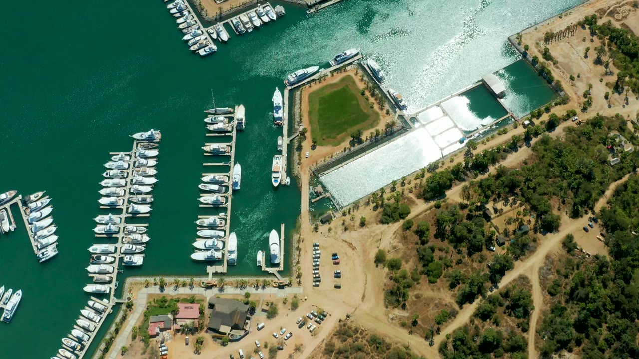 Overhead view of Puerto Los Cabos marina at the start of the clip, with docked white boats on green water, sandy lots, trees, parked cars, and no visible people.