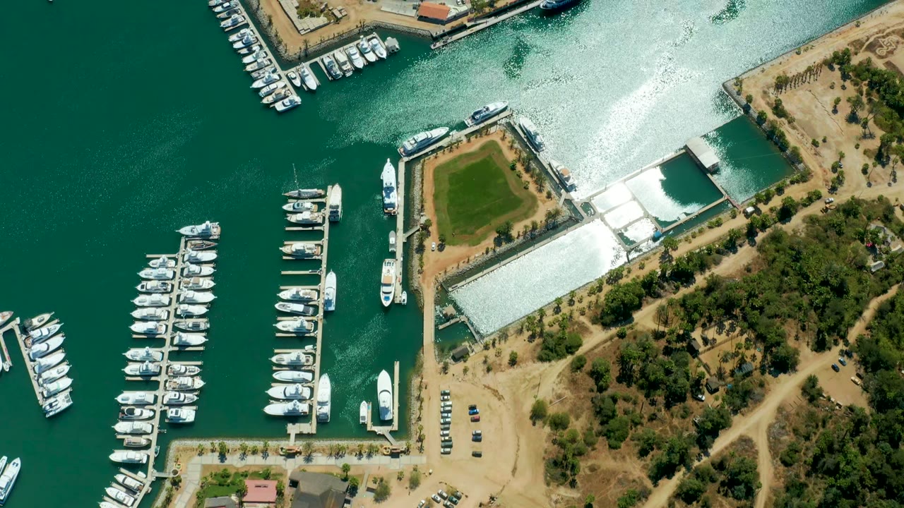 Top-down view of Puerto Los Cabos marina two seconds in, with more open green water at left, docked boats along piers, sandy ground, tree cover, and no visible people.