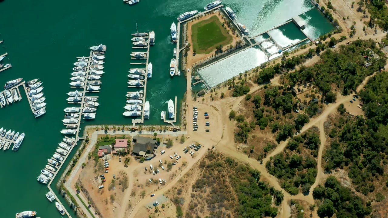 Opening overhead view of Puerto Los Cabos marina with rows of docked white boats, green-blue water, dry paths, low buildings, trees, and no visible people.