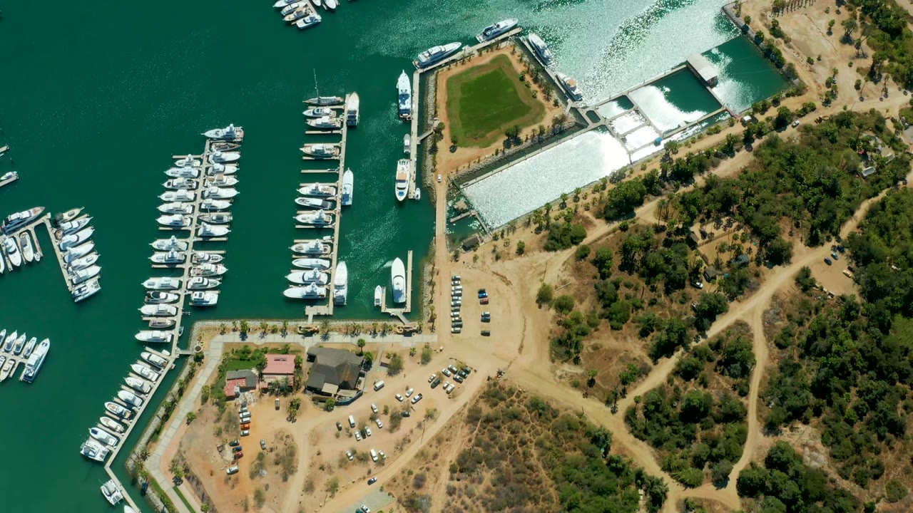 At two seconds, the Puerto Los Cabos marina remains in a high overhead view with bright water reflection at the top, boats lining the docks, tan roads, trees, and no visible people.
