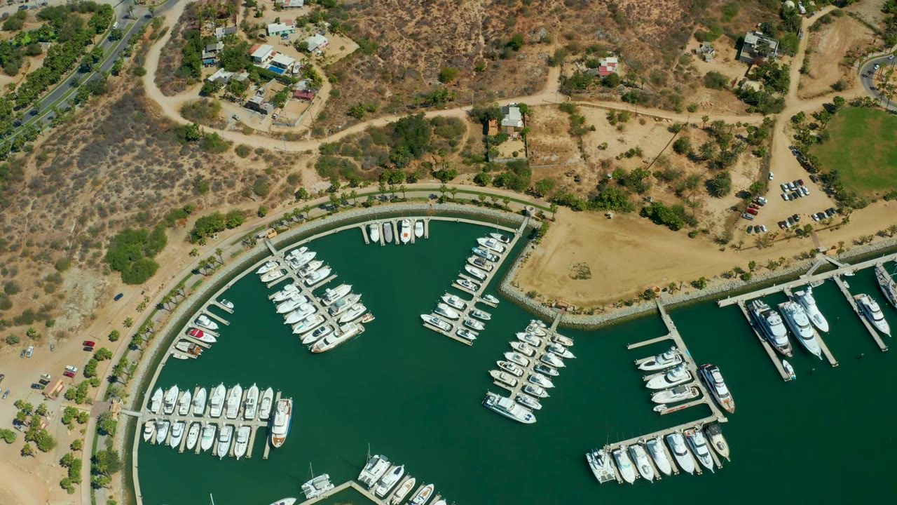 Opening aerial frame over Puerto Los Cabos marina, with white docked boats in dark green water, curved docks, dry hillside terrain, scattered buildings, roads, cars, and no visible people.