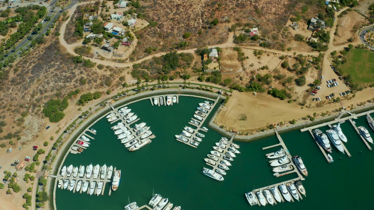 Aerial frame of the Puerto Los Cabos marina basin with dense rows of docked boats below, a curved seawall, dry lots, hillside brush, road bends, vehicles, and no visible people.