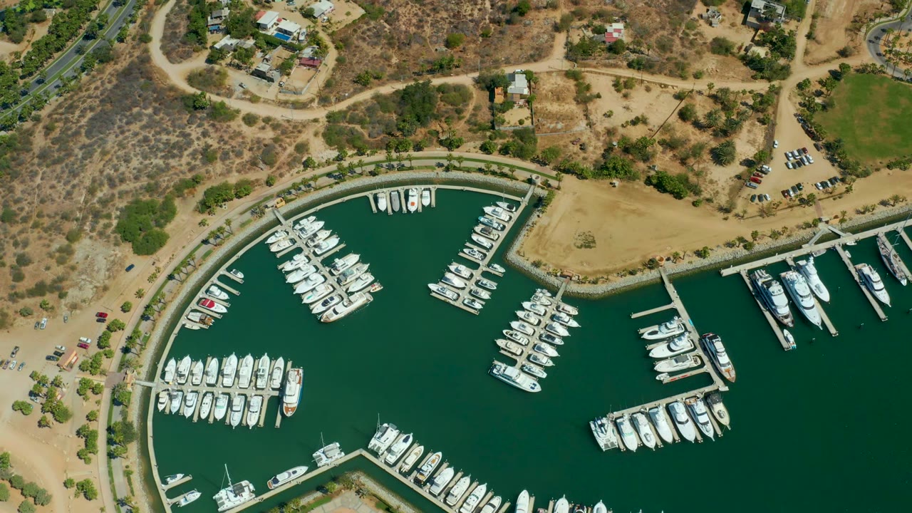 Overhead 4K view of Puerto Los Cabos marina with rows of white boats in a curved green basin, dry land and roads above the harbor, and no visible people.