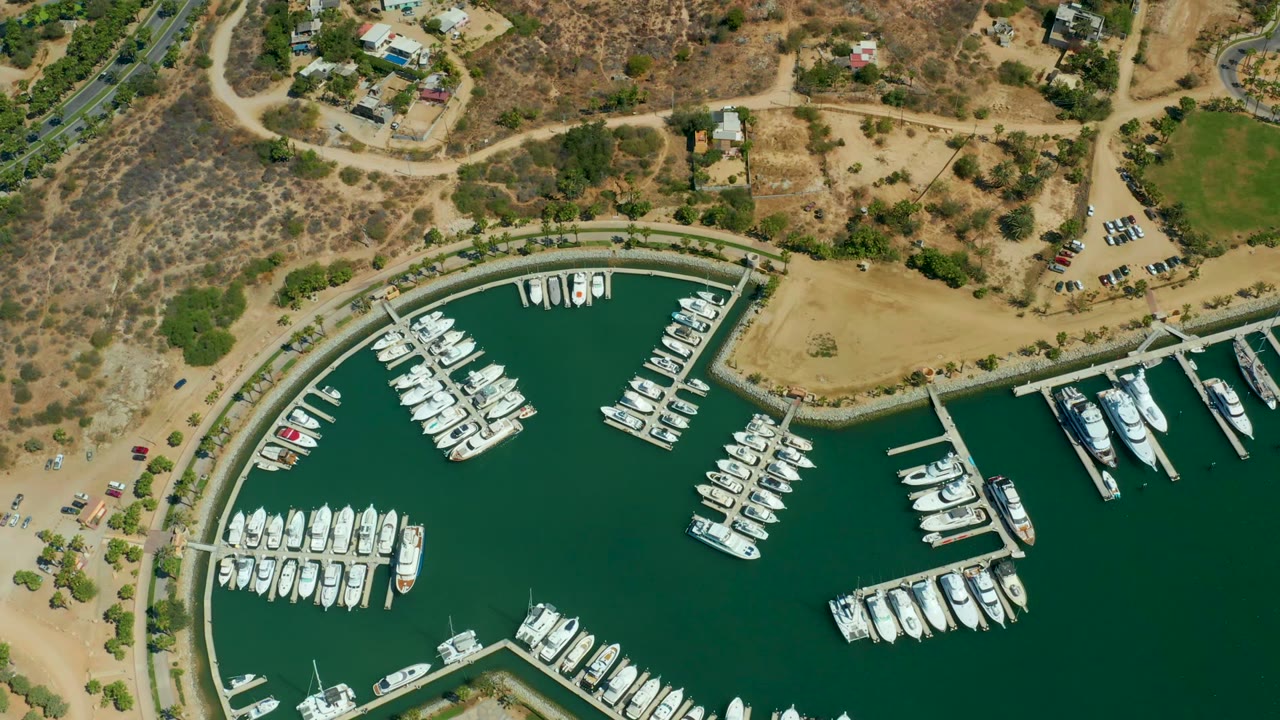 Aerial marina basin with white boats docked along piers and dry land beyond the shore.